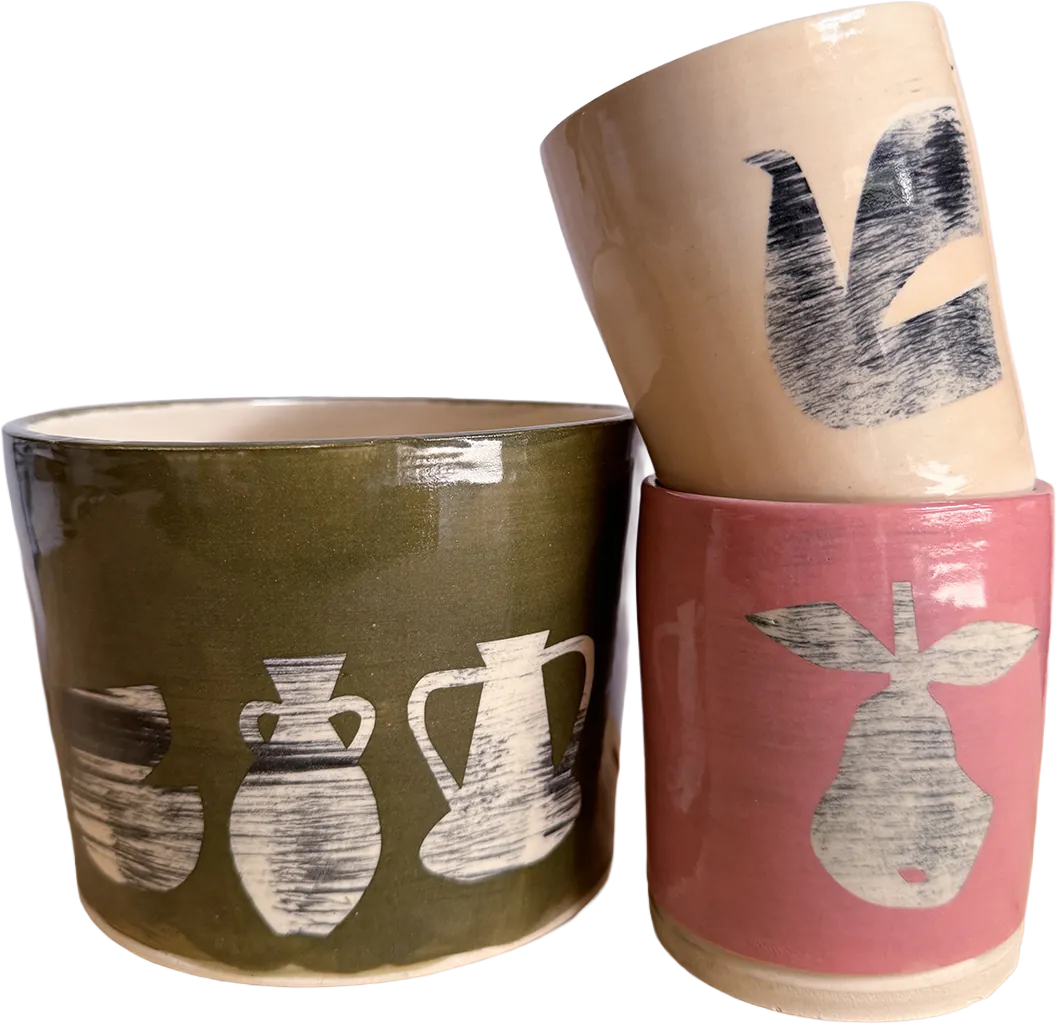 Three vessels: an olive cup painted with white amphora shapes, a coral cup with a pear, and an off-white cup with brushed black marks in the shape of a dove.